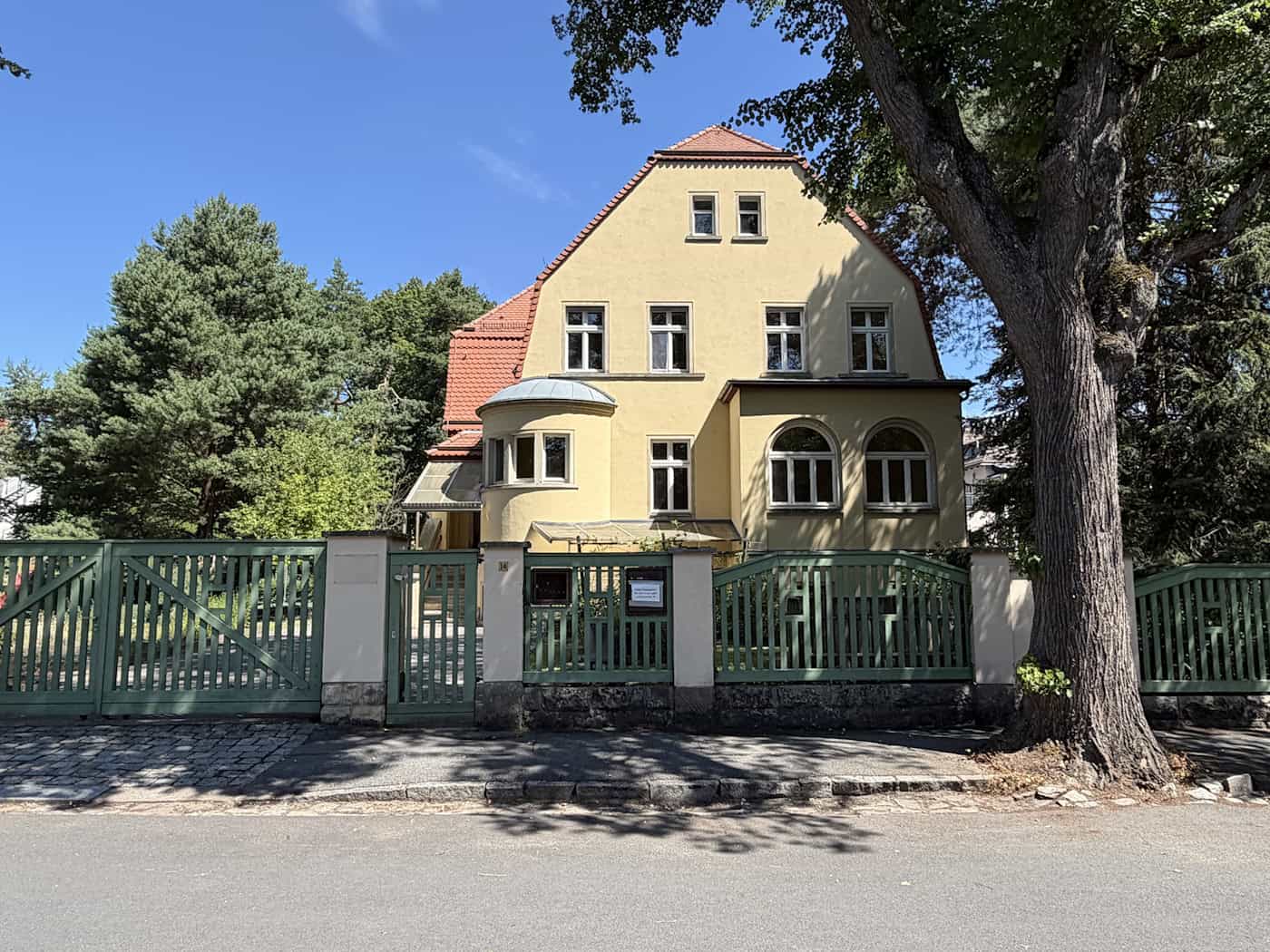 Villa Darwinstraße 14 in Dresden-Klotzsche: Denkmal im Reformstil zu verkaufen Gelb verputzte Villa im Reformstil mit grünem Holzzaun, Eckpavillon und großem Laubbaum im Vordergrund. Das denkmalgeschützte Anwesen in der Darwinstraße steht in einem begrünten Wohngebiet unter blauem Himmel.