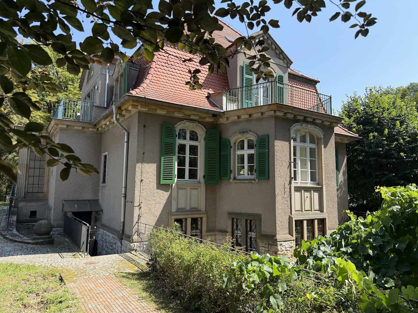 Denkmalgeschützte Villa am Dörnichtweg 32 in Dresden-Klotzsche zu verkaufen Neuklassizistische Villa mit grauer Putzfassade, grünen Klappläden, Dachbalkon und Walmdach. Das Gebäude ist von dichtem Grün und Bäumen umgeben, ein gepflasterter Weg führt entlang des Hangs zum Souterrain.