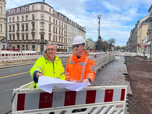 Steffen Lohmann (links) in gelber Warnjacke und Alexander Richter mit weißem Bauhelm blicken gemeinsam hinter einer rot-weißen Absperrung in einen Bauplan. Im Hintergrund erstreckt sich die Königsbrücker Straße in Dresden mit Häuserfassaden und Bohrturm.