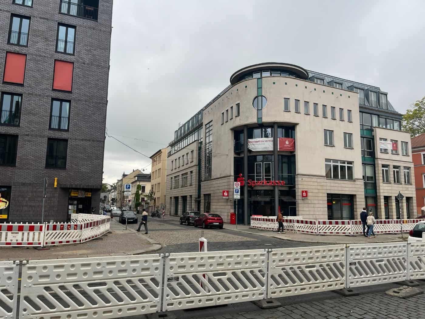 Straßenansicht an der Ecke Königsbrücker Straße und Katharinenstraße in Dresden mit einem modernen Sparkassengebäude im Hintergrund. Im Vordergrund stehen weiß-rote Baustellen-Sicherheitszäune auf der Fahrbahn.
