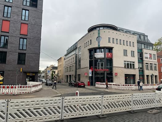 Straßenansicht an der Ecke Königsbrücker Straße und Katharinenstraße in Dresden mit einem modernen Sparkassengebäude im Hintergrund. Im Vordergrund stehen weiß-rote Baustellen-Sicherheitszäune auf der Fahrbahn.
