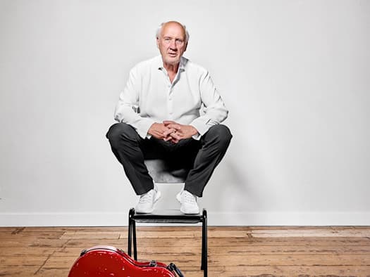 Der niederländische Künstler Herman van Veen sitzt in weißem Hemd mit verschränkten Händen auf einem Stuhl im Studio. Vor ihm auf dem Holzboden liegt ein roter Instrumentenkoffer mit einem Aufkleber mit der Aufschrift „TOUR 2027/28“. (Foto: William Rutten)