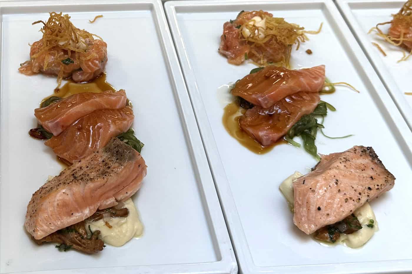 Dreierlei vom Lachs beim Charity-Kochen für Gastronomie-Nachwuchs in Dresden Weiße, rechteckige Porzellanteller mit drei feinen Lachs-Variationen: Lachs-Tatar mit knusprigem Topping, marinierte Lachsstreifen und sanft gebratenes Lachsfilet auf hellem Püree.