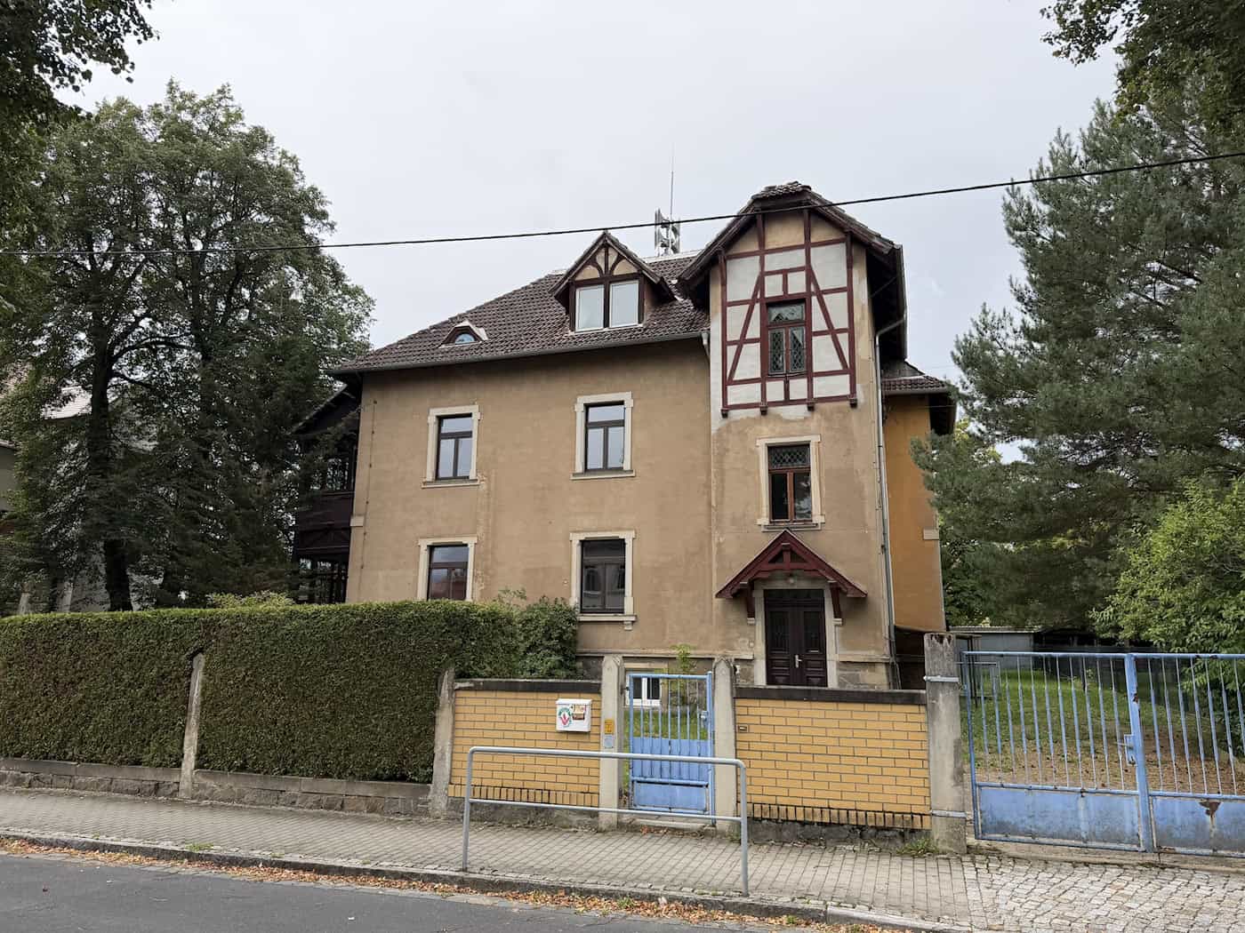 Jugendstil-Villa in Dresden-Langebrück: Jakob-Weinheimer-Straße 16 steht zum Verkauf Freistehendes, zweigeschossiges Wohnhaus mit Jugendstilelementen und prägnantem Zierfachwerk am Giebel. Im Vordergrund verläuft eine gelbe Klinkermauer mit blauem Metalltor vor einer dichten Hecke.