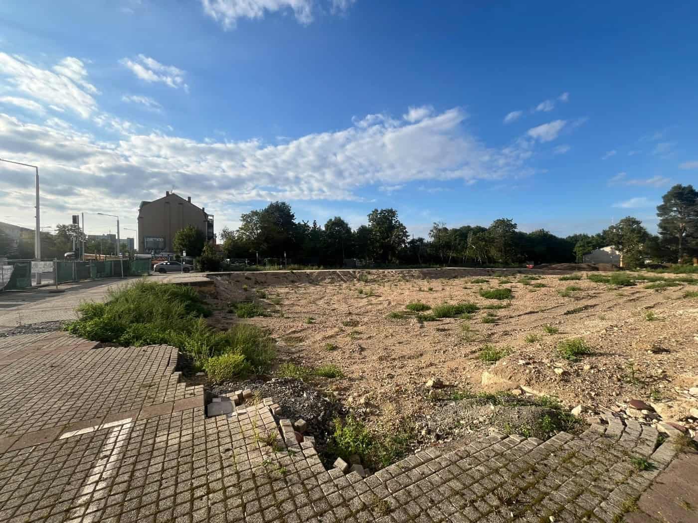 Eine große, beräumte Sandfläche mit vereinzeltem Grün unter blauem Himmel. Im Vordergrund gepflasterter Boden, im Hintergrund Bäume und Gebäude. Es handelt sich um das Bauareal der ehemaligen Operette in Dresden-Leuben.