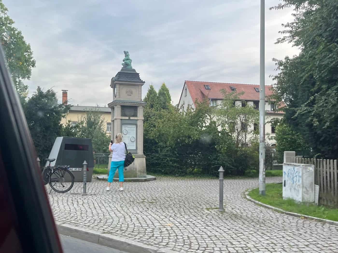 Straßenansicht in Dresden-Lockwitz: Neben einer historischen Wettersäule mit Froschfigur steht ein dunkler Blitzer-Anhänger auf dem Pflaster. Eine Frau steht davor, im Hintergrund befinden sich Bäume und Wohnhäuser.