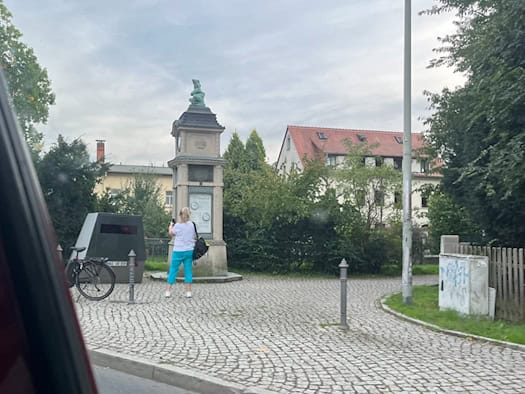 Straßenansicht in Dresden-Lockwitz: Neben einer historischen Wettersäule mit Froschfigur steht ein dunkler Blitzer-Anhänger auf dem Pflaster. Eine Frau steht davor, im Hintergrund befinden sich Bäume und Wohnhäuser.