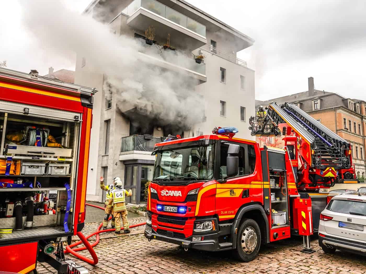 Ein rotes Drehleiterfahrzeug der Feuerwehr steht vor einem modernen Wohnhaus in Dresden-Löbtau, aus dessen zerstörtem Balkonfenster im ersten Stock dichter grauer Rauch quillt. Einsatzkräfte in Schutzkleidung befinden sich am Boden und an der Drehleiter.