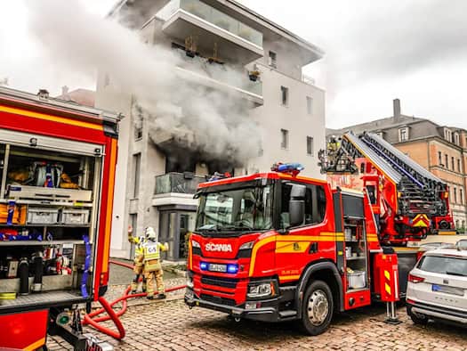 Ein rotes Drehleiterfahrzeug der Feuerwehr steht vor einem modernen Wohnhaus in Dresden-Löbtau, aus dessen zerstörtem Balkonfenster im ersten Stock dichter grauer Rauch quillt. Einsatzkräfte in Schutzkleidung befinden sich am Boden und an der Drehleiter.