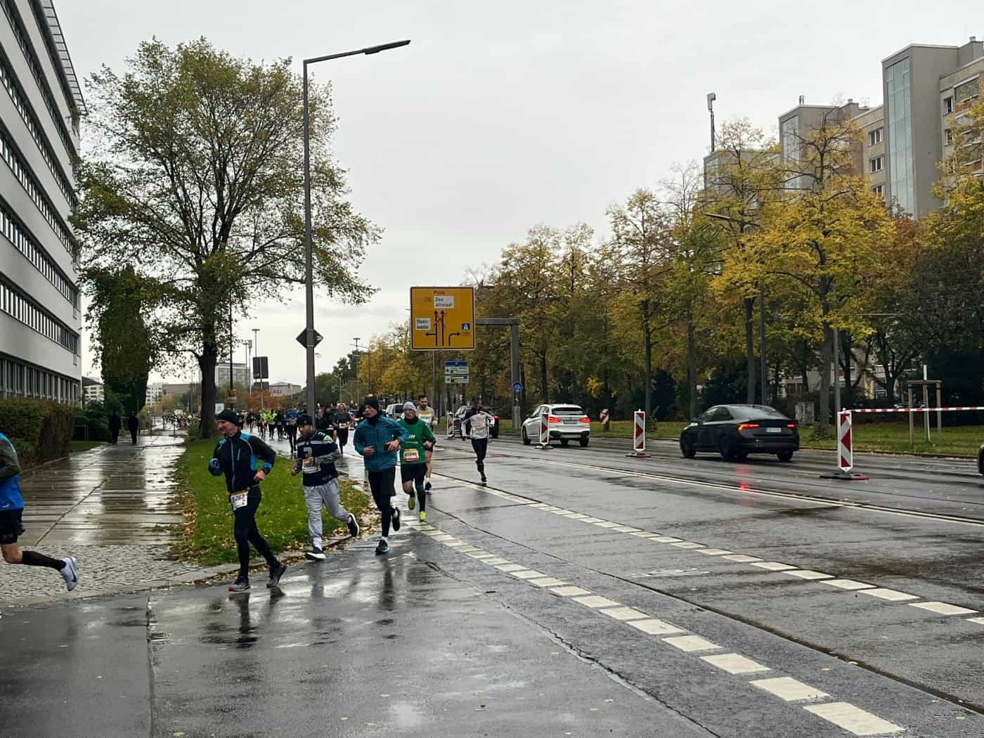 Zahlreiche Läuferinnen und Läufer in Sportbekleidung und mit Startnummern bewegen sich bei nassem Wetter auf einer gesperrten Straße fort. Am Straßenrand stehen herbstliche Bäume, ein gelbes Verkehrsschild und Autos im Hintergrund.