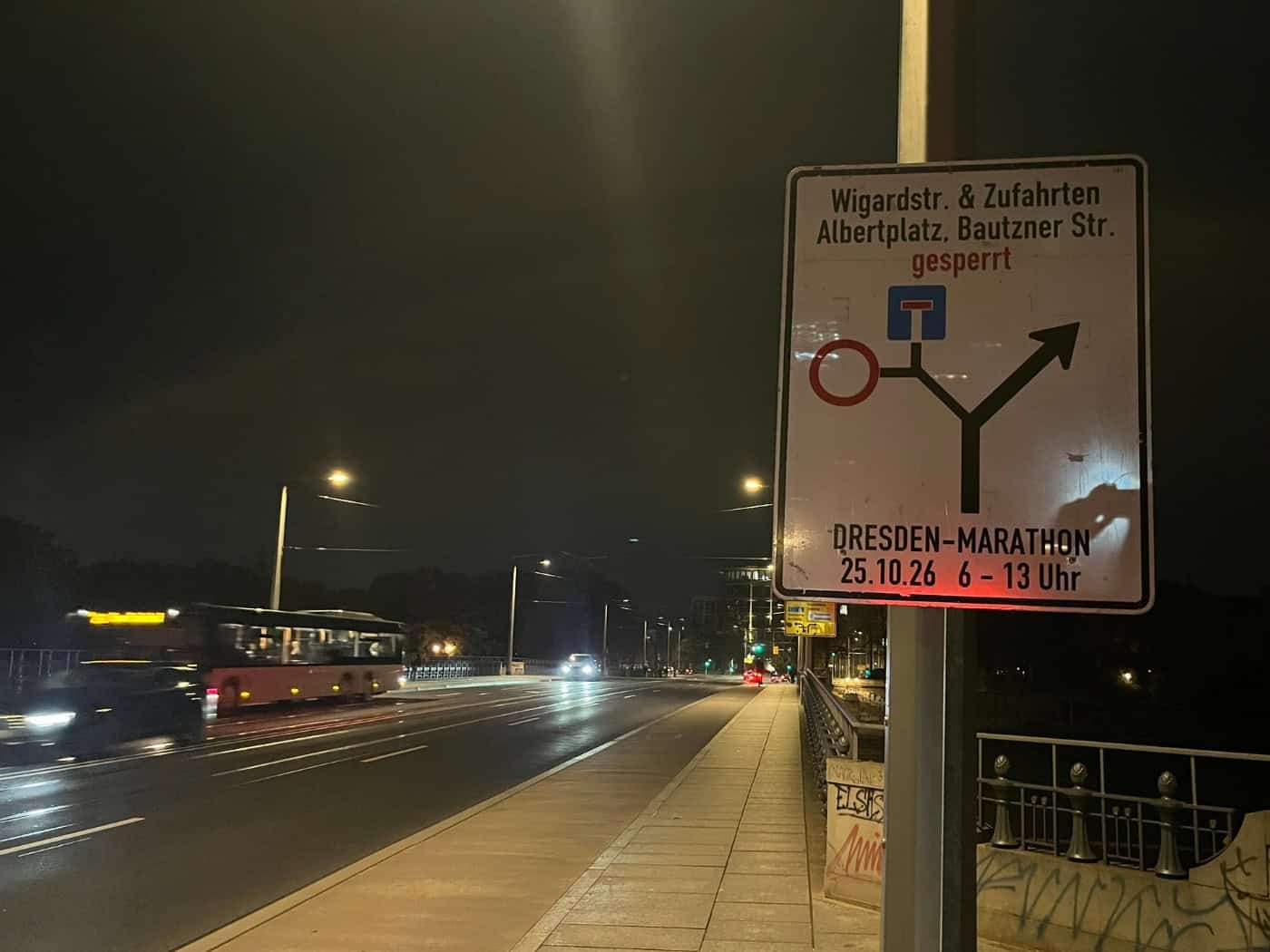 Nachtaufnahme eines weißen Hinweisschildes an einem Laternenmast zur Sperrung der Wigardstraße und Zufahrten zum Albertplatz für den Dresden-Marathon am 25.10.26. Im Hintergrund führt eine beleuchtete Brücke mit vorbeifahrendem Bus durch die Nacht.