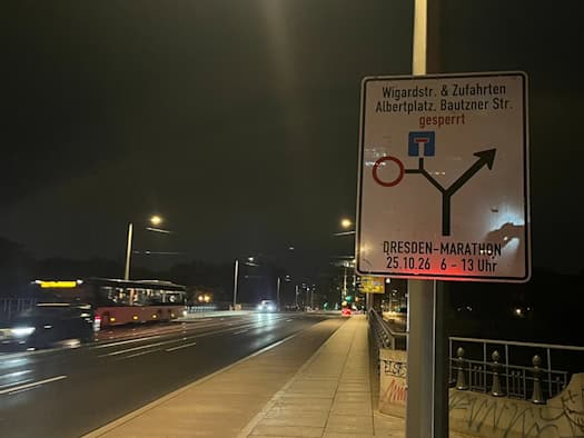 Nachtaufnahme eines weißen Hinweisschildes an einem Laternenmast zur Sperrung der Wigardstraße und Zufahrten zum Albertplatz für den Dresden-Marathon am 25.10.26. Im Hintergrund führt eine beleuchtete Brücke mit vorbeifahrendem Bus durch die Nacht.
