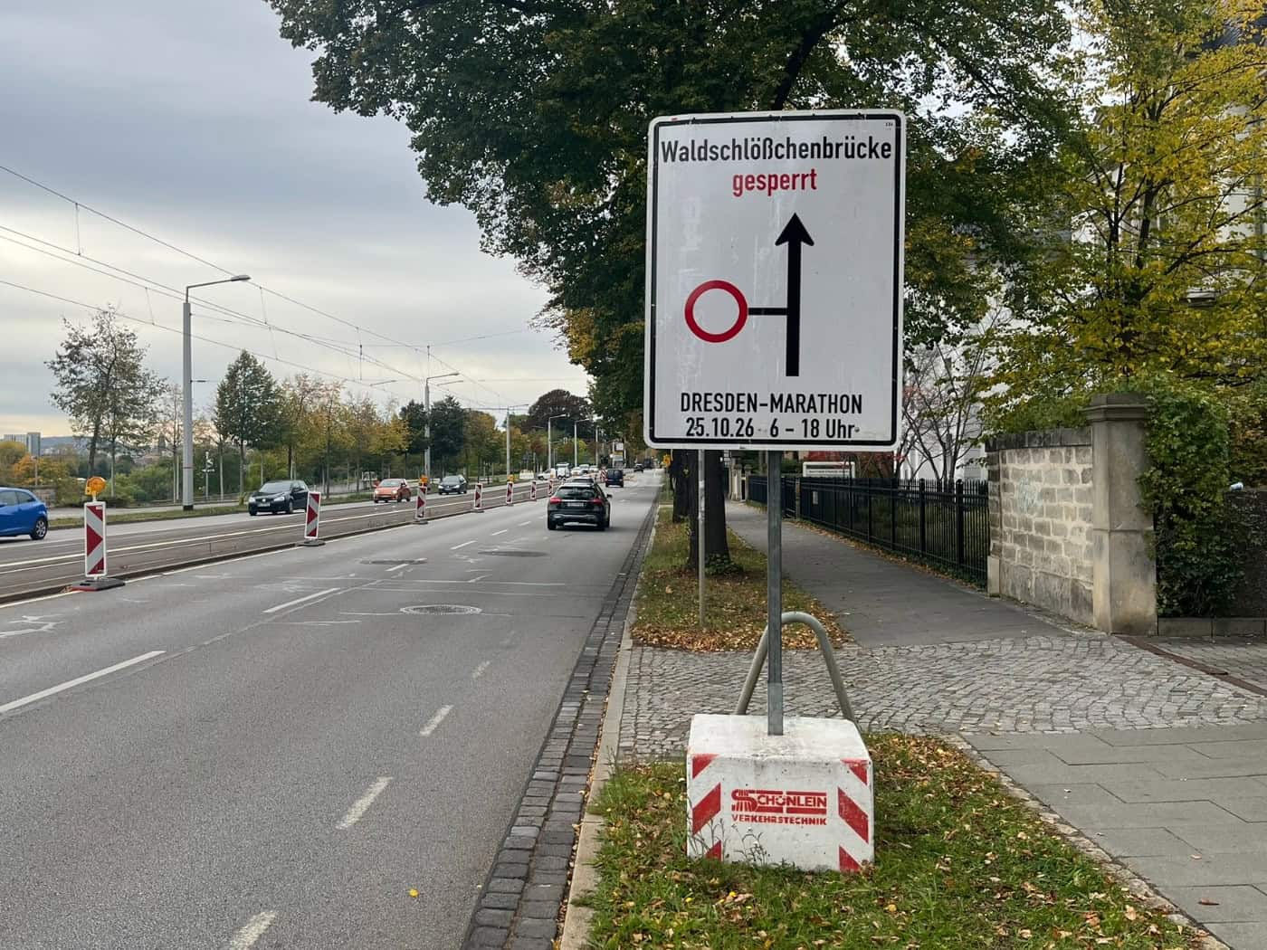 Ein weißes Verkehrsschild am Straßenrand warnt vor der Sperrung der Waldschlößchenbrücke für den Dresden-Marathon am 25.10.26 von 6 bis 18 Uhr. Im Hintergrund verläuft eine mehrspurige Straße mit fahrenden Autos und herbstlichen Bäumen.