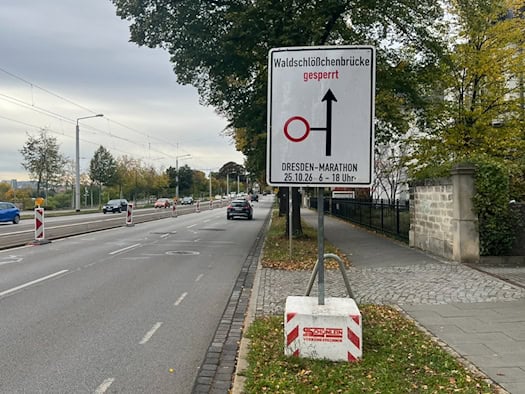 Ein weißes Verkehrsschild am Straßenrand warnt vor der Sperrung der Waldschlößchenbrücke für den Dresden-Marathon am 25.10.26 von 6 bis 18 Uhr. Im Hintergrund verläuft eine mehrspurige Straße mit fahrenden Autos und herbstlichen Bäumen.