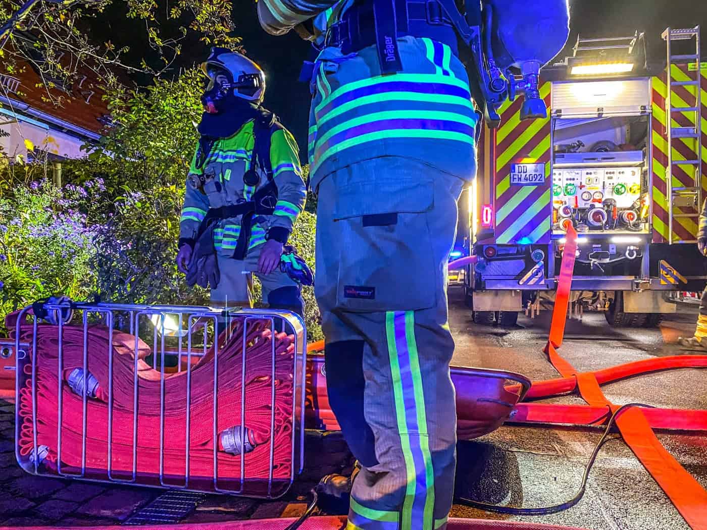 Feuerwehrleute in reflektierender Schutzkleidung bei einem nächtlichen Einsatz: Im Vordergrund liegt ein metallener Schlauchtragekorb mit roten Schläuchen, im Hintergrund ist das beleuchtete Heck eines Feuerwehrfahrzeugs zu sehen.
