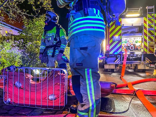 Feuerwehrleute in reflektierender Schutzkleidung bei einem nächtlichen Einsatz: Im Vordergrund liegt ein metallener Schlauchtragekorb mit roten Schläuchen, im Hintergrund ist das beleuchtete Heck eines Feuerwehrfahrzeugs zu sehen.