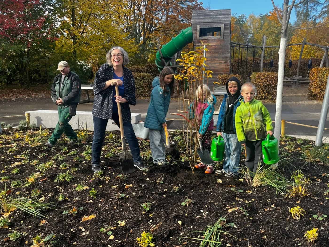 Umweltbürgermeisterin Eva Jähnigen steht lächelnd mit Spaten im frisch angelegten Beet der Montessorischule Dresden. Neben ihr setzen Grundschulkinder mit Schaufeln und grünen Gießkannen eine junge Felsenbirne in die Erde.