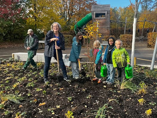 Umweltbürgermeisterin Eva Jähnigen steht lächelnd mit Spaten im frisch angelegten Beet der Montessorischule Dresden. Neben ihr setzen Grundschulkinder mit Schaufeln und grünen Gießkannen eine junge Felsenbirne in die Erde.