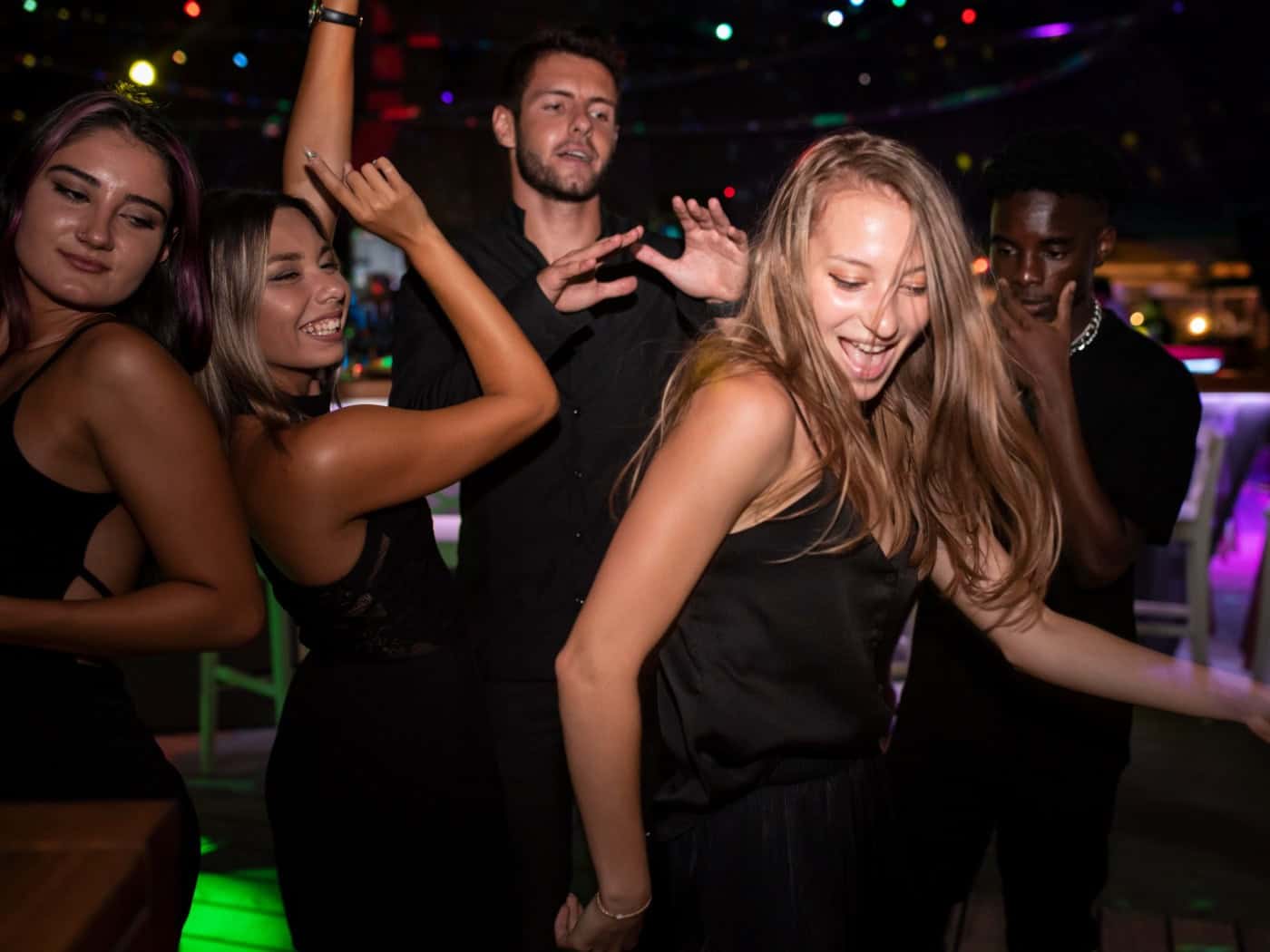 Eine Gruppe junger Erwachsener tanzt ausgelassen in einem abgedunkelten Club. Im Vordergrund tanzt eine blonde Frau in einem schwarzen Top mit schwingendem Haar, umgeben von lächelnden Freunden unter farbigen Partylichtern.