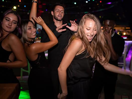 Eine Gruppe junger Erwachsener tanzt ausgelassen in einem abgedunkelten Club. Im Vordergrund tanzt eine blonde Frau in einem schwarzen Top mit schwingendem Haar, umgeben von lächelnden Freunden unter farbigen Partylichtern.