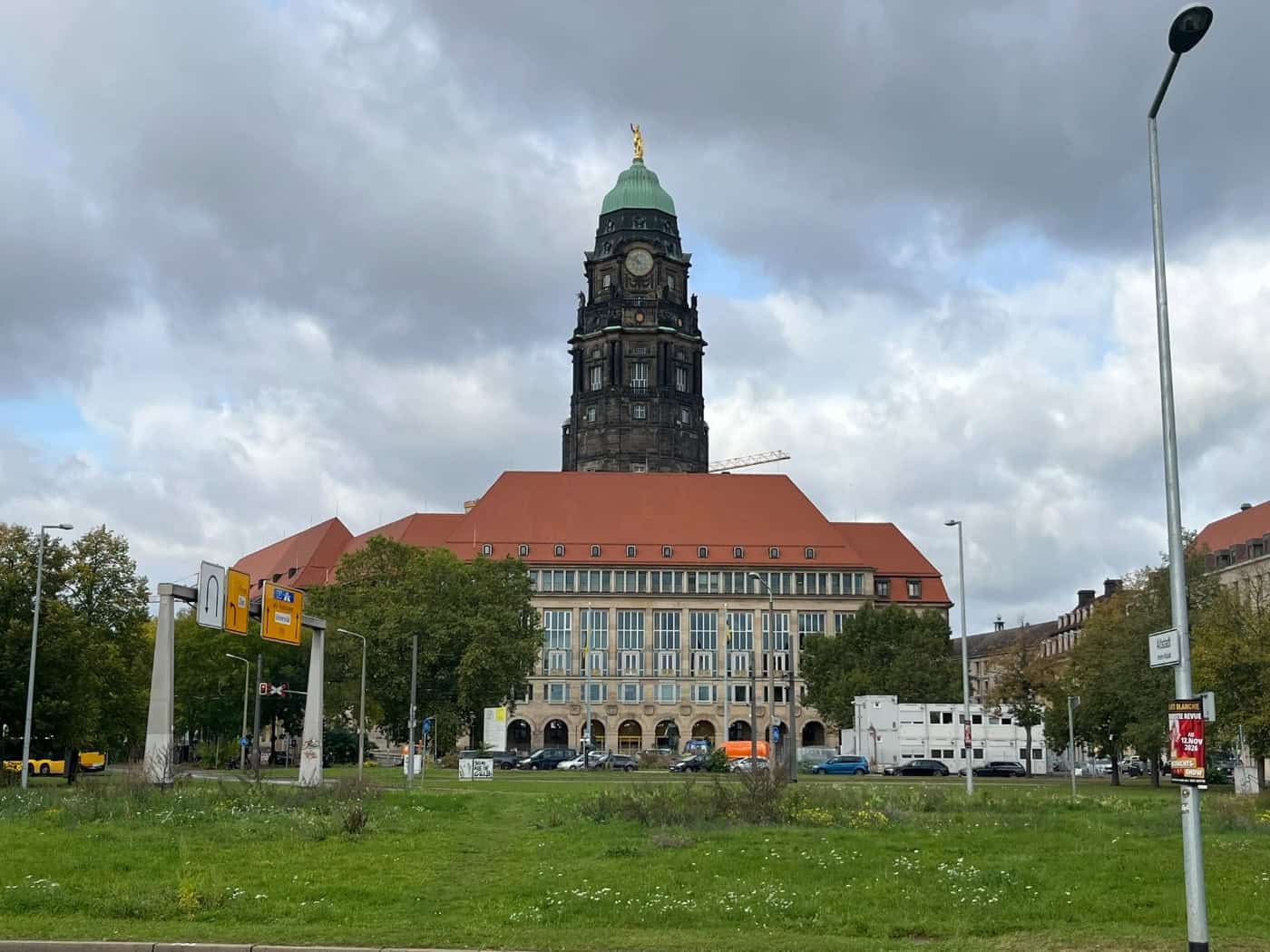 Außenaufnahme des Neuen Rathauses in Dresden mit seinem prägnanten Rathausturm und der goldenen St.-Benno-Statue an der Spitze. Im Vordergrund liegen eine grüne Rasenfläche, Straßenbeschilderungen sowie Laternenmasten unter leicht bewölktem Himmel.
