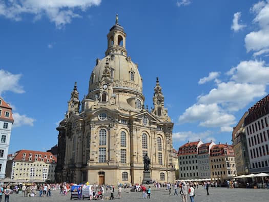 Totale Aufnahme des belebten Neumarkts in Dresden bei Sonnenschein. In der Bildmitte steht die imposante Frauenkirche mit ihrer großen Kuppel, davor die Statue von Martin Luther. Menschen spazieren über den gepflasterten Platz.