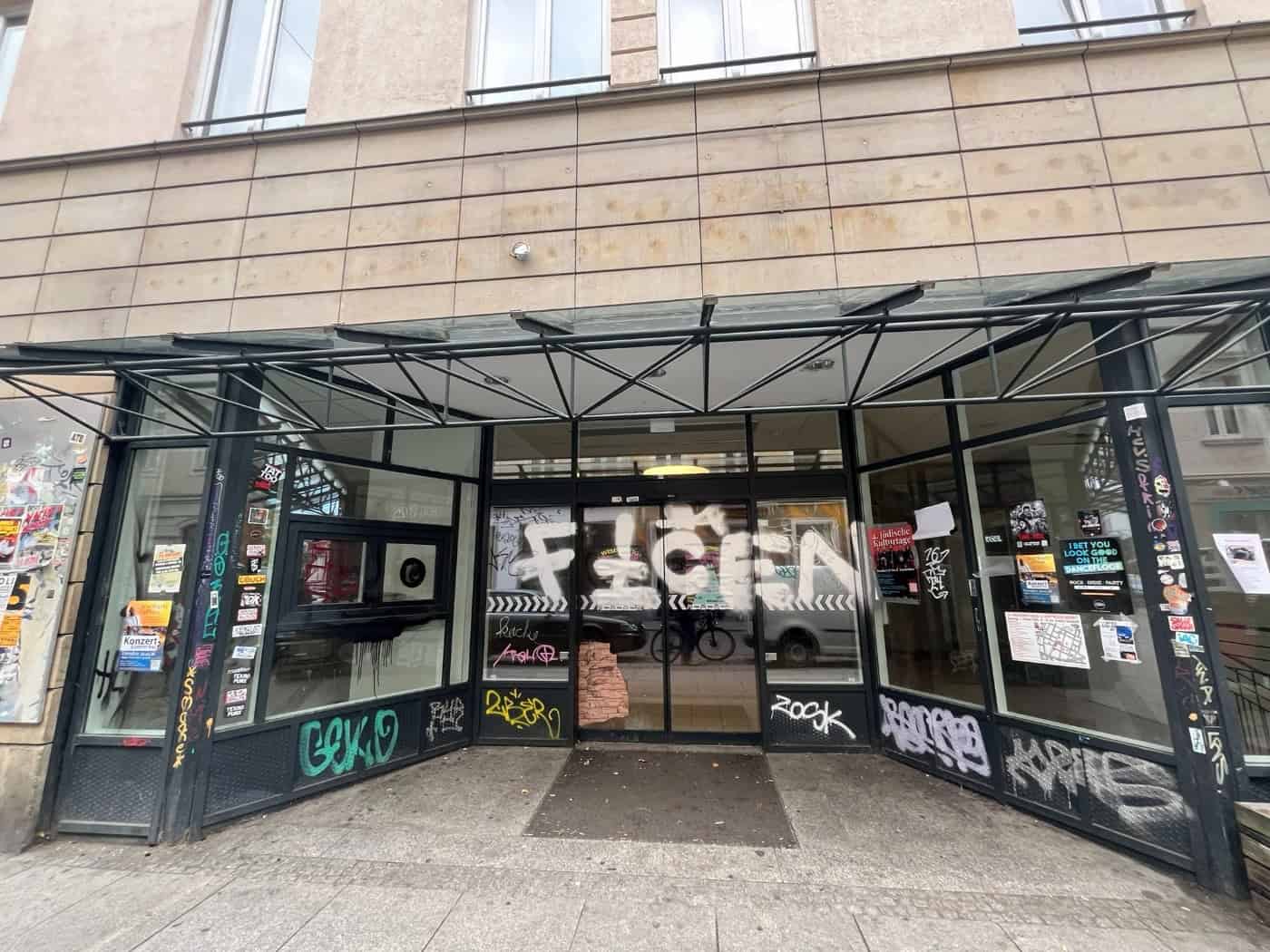 Frontansicht einer leerstehenden Ladeneinheit an der Alaunstraße in Dresden mit gläsernem Vordach. Die Scheiben und Schiebetüren des ehemaligen Konsum-Supermarktes sind mit Graffiti und Aufklebern versehen, an den Wänden hängen Veranstaltungsplakate.