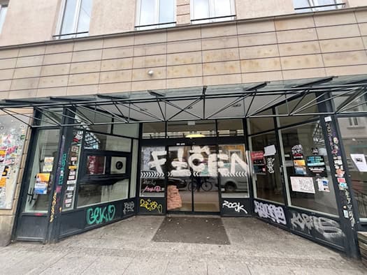 Frontansicht einer leerstehenden Ladeneinheit an der Alaunstraße in Dresden mit gläsernem Vordach. Die Scheiben und Schiebetüren des ehemaligen Konsum-Supermarktes sind mit Graffiti und Aufklebern versehen, an den Wänden hängen Veranstaltungsplakate.