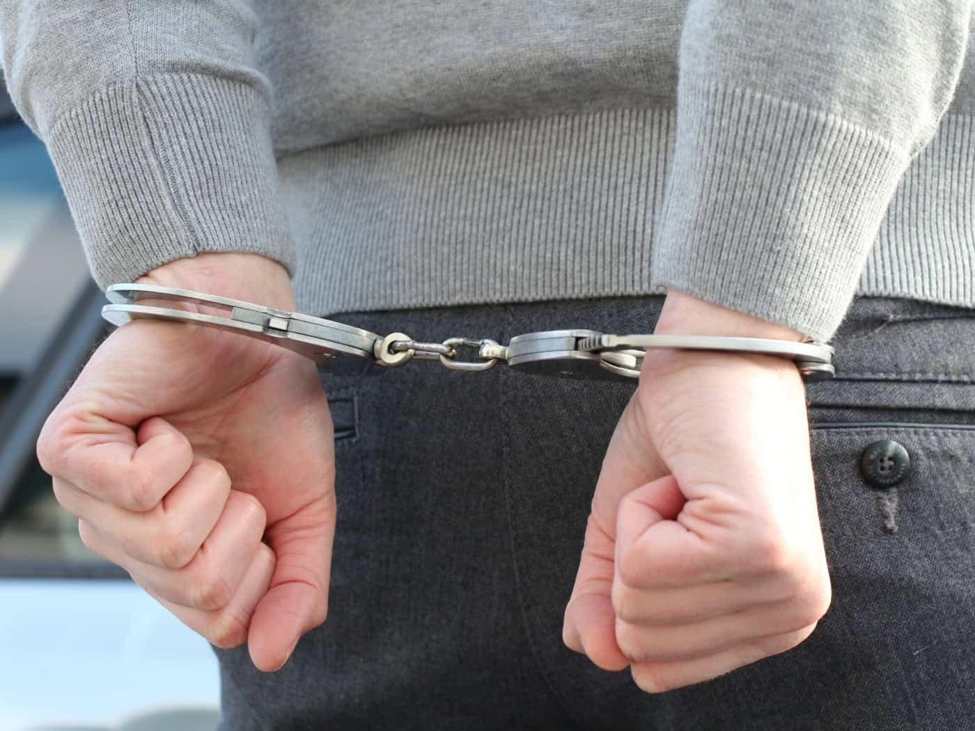 Symbolbild: Nahaufnahme der Hände eines Tatverdächtigen in Handschellen hinter dem Rücken. Er trägt einen grauen Pullover und eine dunkle Hose, im Hintergrund ist ein Polizeifahrzeug angedeutet.