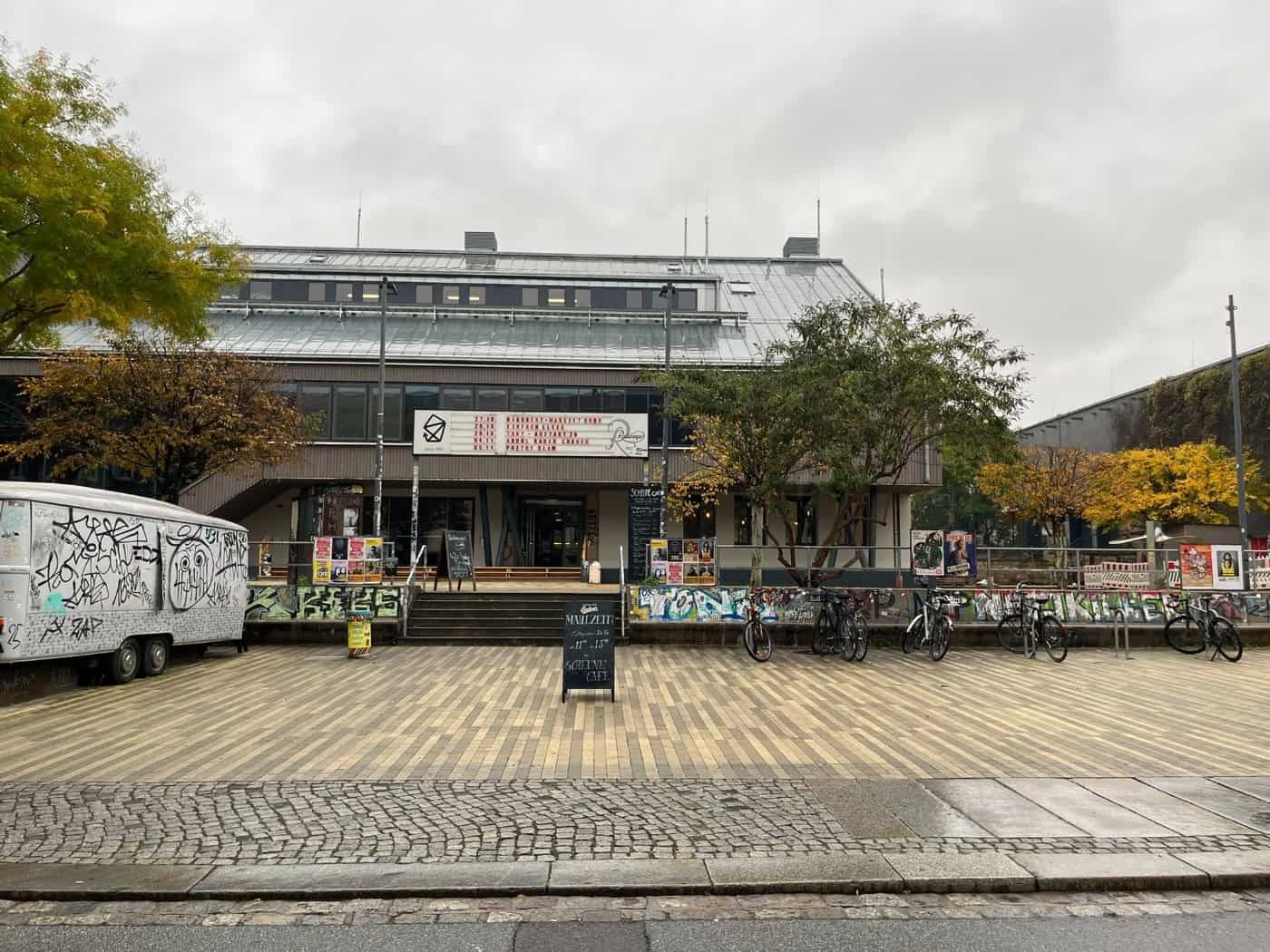 Frontalansicht des gepflasterten Scheune-Vorplatzes in der Dresdner Äußeren Neustadt. Im Hintergrund steht das Kulturzentrum Scheune mit Treppenaufgang, links parkt ein graffitibeschmierter Anhänger, davor stehen Fahrräder und ein Aufsteller unter bewölktem Himmel.