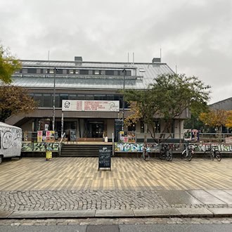 Frontalansicht des gepflasterten Scheune-Vorplatzes in der Dresdner Äußeren Neustadt. Im Hintergrund steht das Kulturzentrum Scheune mit Treppenaufgang, links parkt ein graffitibeschmierter Anhänger, davor stehen Fahrräder und ein Aufsteller unter bewölktem Himmel.