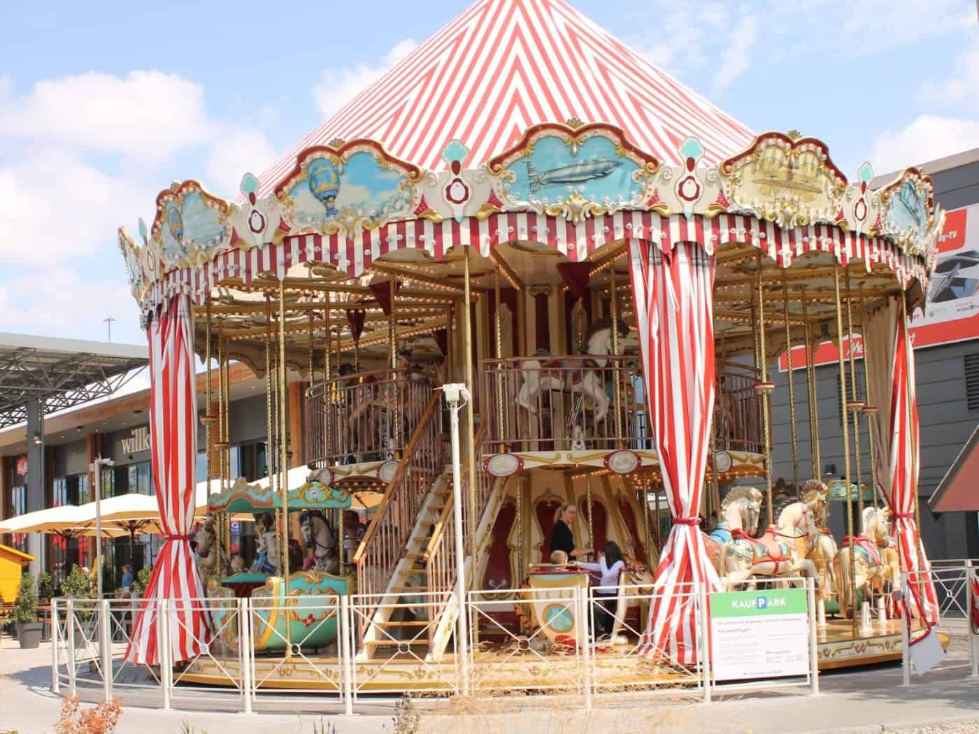 Prachtvolles, zweistöckiges historisches Karussell mit rot-weiß gestreiftem Zeltdach und reich verzierten Pferdefiguren vor dem Kaufpark Dresden-Nickern. Das Fahrgeschäft steht auf einer Pflasterfläche vor modernen Einkaufspassagen unter sonnigem Himmel.