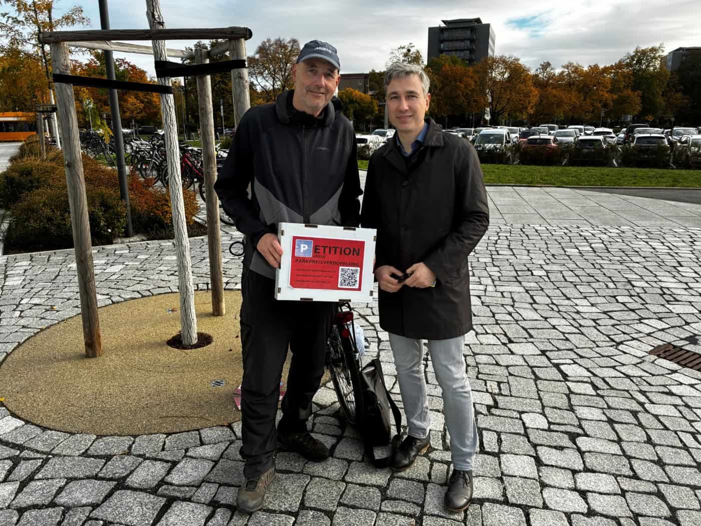 Petent Torsten Küllig (links) mit Kappe und Schild zur E-Petition gegen Parkpreisverdopplung steht neben Dresdens Baubürgermeister Stephan Kühn (rechts im dunklen Mantel) auf dem gepflasterten Vorplatz des Stadtforums, im Hintergrund parkende Autos.
