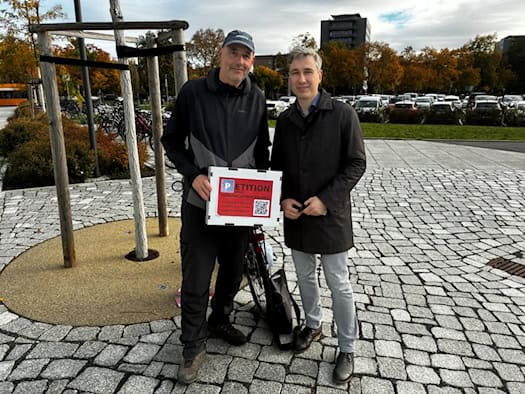 Petent Torsten Küllig (links) mit Kappe und Schild zur E-Petition gegen Parkpreisverdopplung steht neben Dresdens Baubürgermeister Stephan Kühn (rechts im dunklen Mantel) auf dem gepflasterten Vorplatz des Stadtforums, im Hintergrund parkende Autos.