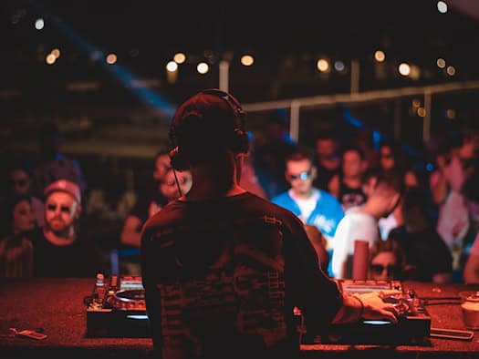 DJ von hinten mit Kopfhörern an den Reglern des Mischpults im Scheinwerferlicht. Im Hintergrund ist die feiernde Menge auf der Tanzfläche eines voll besetzten Clubs zu sehen.