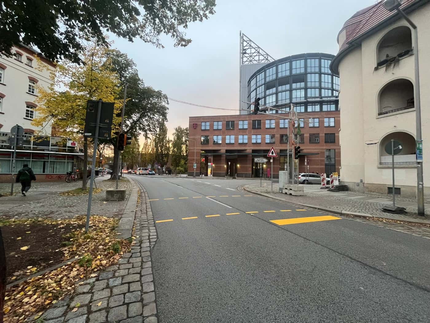 Blick entlang der Riesaer Straße in Dresden-Pieschen mit gelben Übergangsmarkierungen auf dem Asphalt und Ampeln im Vordergrund. Im Hintergrund sind moderne Bürogebäude, herbstliche Bäume und Gehwege zu sehen.