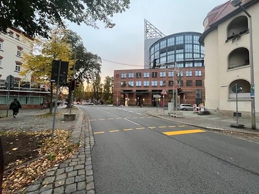 Blick entlang der Riesaer Straße in Dresden-Pieschen mit gelben Übergangsmarkierungen auf dem Asphalt und Ampeln im Vordergrund. Im Hintergrund sind moderne Bürogebäude, herbstliche Bäume und Gehwege zu sehen.