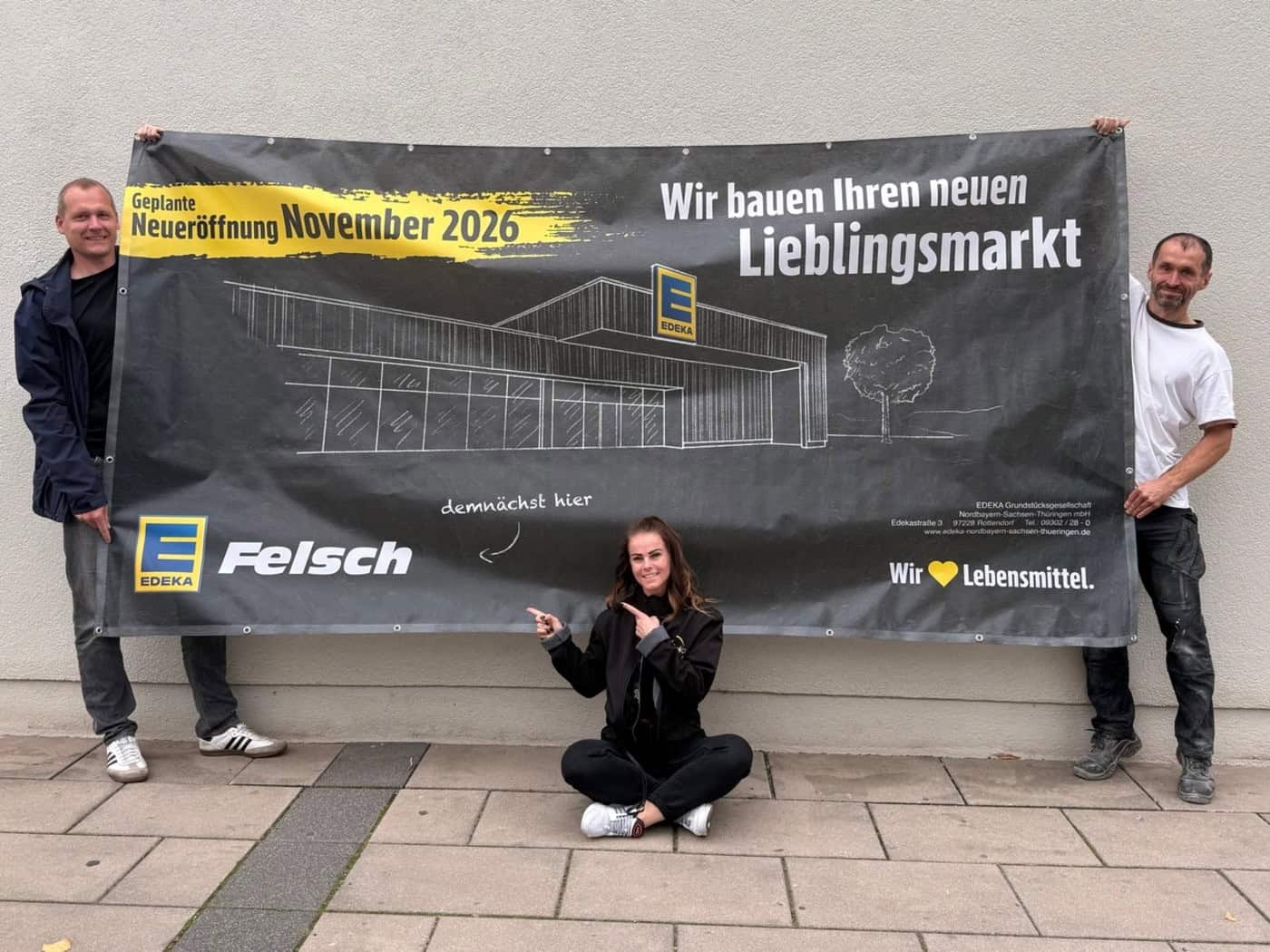 Drei Personen halten ein großes Ankündigungsbanner mit der Aufschrift Edeka Felsch und Neueröffnung November 2026. Die Betreiberin sitzt in der Mitte auf dem Gehweg und zeigt auf den Schriftzug.