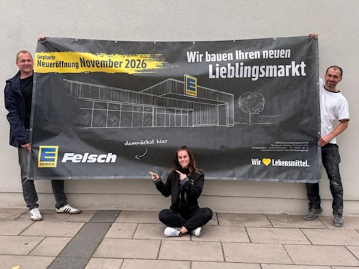 Drei Personen halten ein großes Ankündigungsbanner mit der Aufschrift Edeka Felsch und Neueröffnung November 2026. Die Betreiberin sitzt in der Mitte auf dem Gehweg und zeigt auf den Schriftzug.