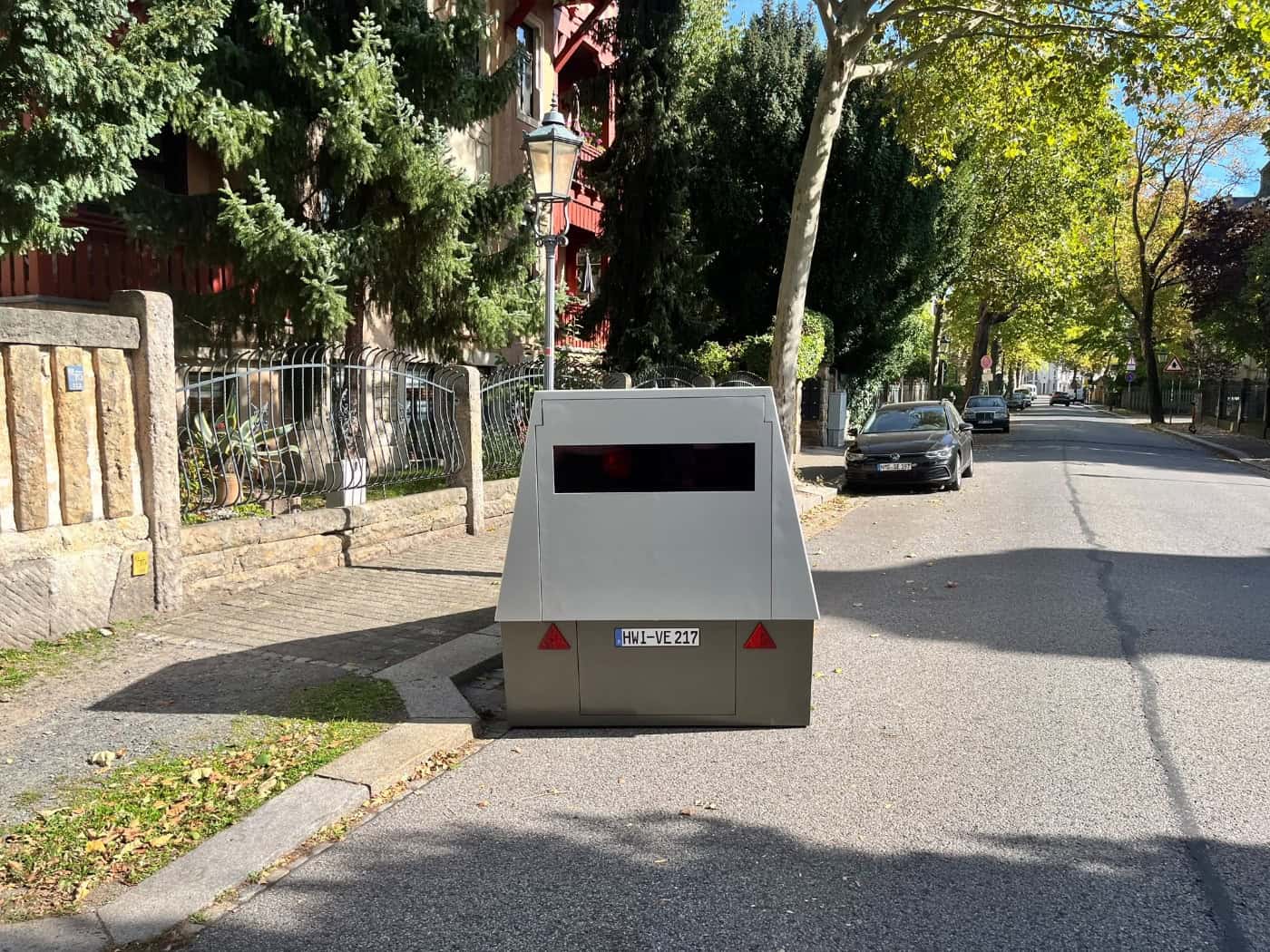 Ein grauer, kastenförmiger Blitzer-Anhänger mit dem Kennzeichen HWI-VE 217 steht am Straßenrand der sonnigen Platanenstraße in Dresden. Im Hintergrund sind Bäume, ein Gehweg und geparkte Autos zu sehen.