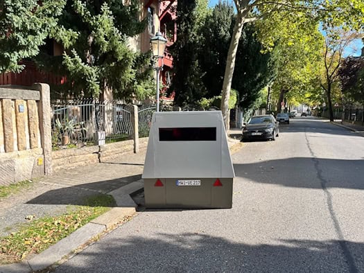 Ein grauer, kastenförmiger Blitzer-Anhänger mit dem Kennzeichen HWI-VE 217 steht am Straßenrand der sonnigen Platanenstraße in Dresden. Im Hintergrund sind Bäume, ein Gehweg und geparkte Autos zu sehen.