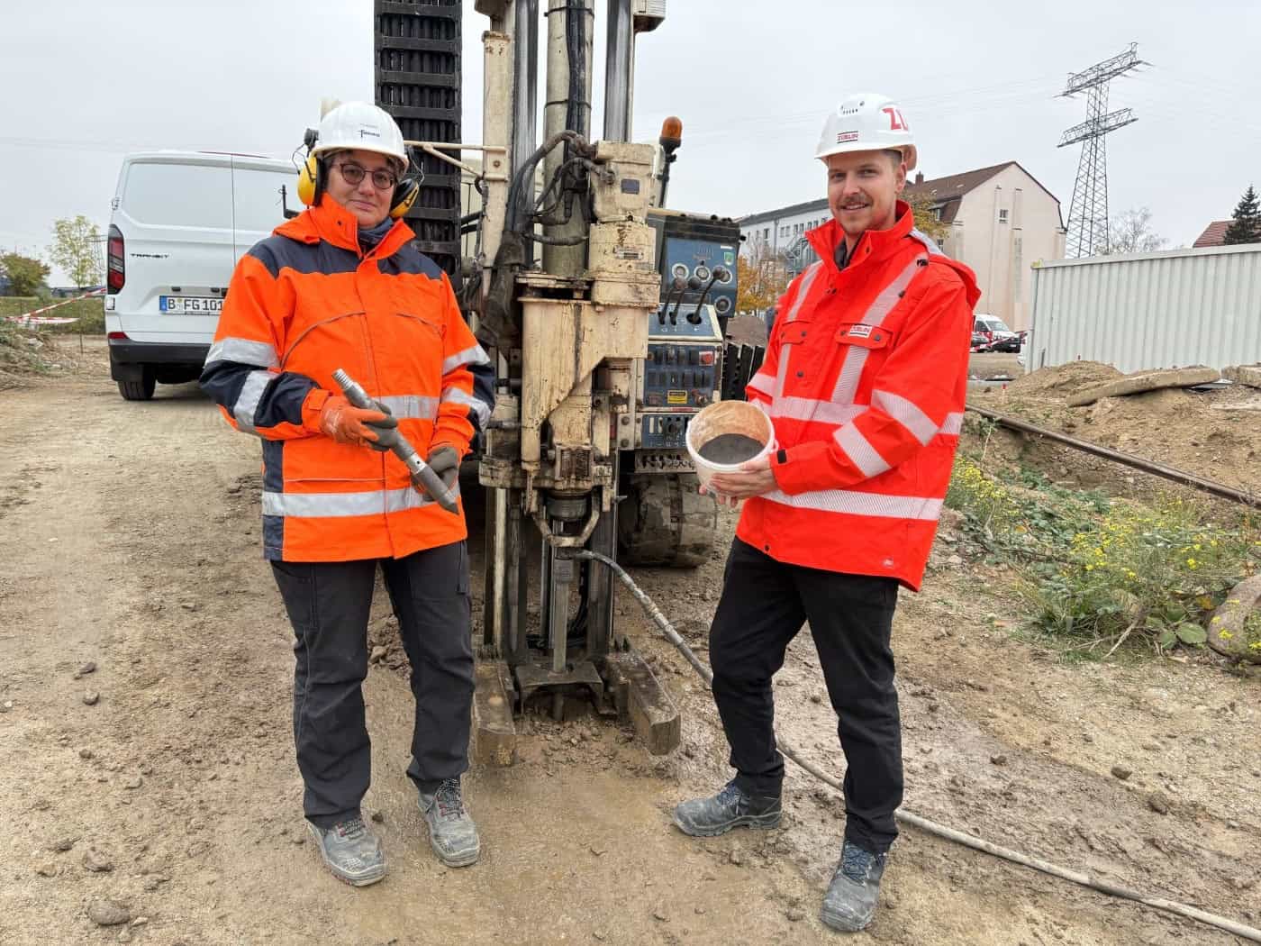 Dr. Christine Laskov in orangefarbener Warnjacke hält eine metallene Injektionslanze. Daneben steht Florian Schramm mit weißem Bauhelm und einem Eimer mit dunklem Eisengranulat-Gemisch vor einem Bohrgerät auf der Baustelle an der Rosenstraße.