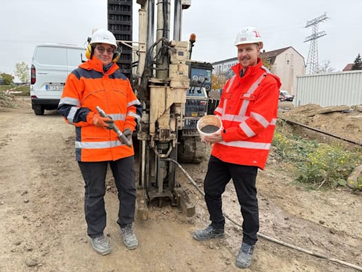 Dr. Christine Laskov in orangefarbener Warnjacke hält eine metallene Injektionslanze. Daneben steht Florian Schramm mit weißem Bauhelm und einem Eimer mit dunklem Eisengranulat-Gemisch vor einem Bohrgerät auf der Baustelle an der Rosenstraße.