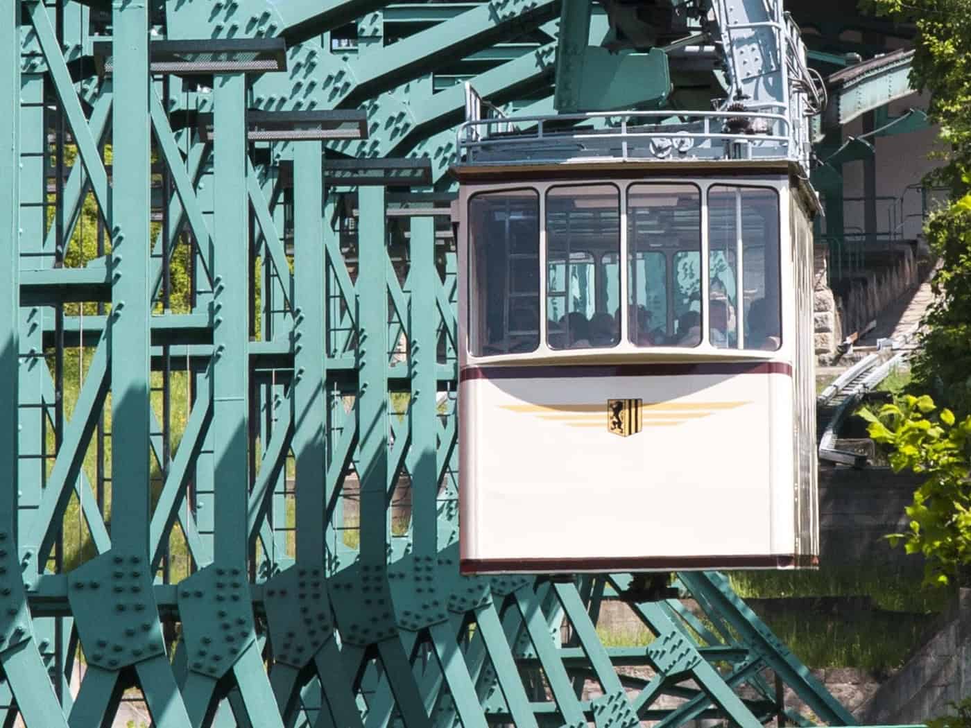Eine beige Gondel der Dresdner Schwebebahn hängt an einem massiven, grün lackierten Stahlgerüst auf dem Weg zur Oberen Station. Im Hintergrund sind grüne Bäume und Menschen auf der Aussichtsplattform der Station unter blauem Himmel zu sehen.