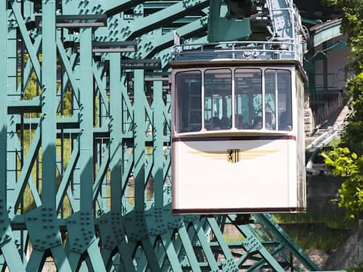 Eine beige Gondel der Dresdner Schwebebahn hängt an einem massiven, grün lackierten Stahlgerüst auf dem Weg zur Oberen Station. Im Hintergrund sind grüne Bäume und Menschen auf der Aussichtsplattform der Station unter blauem Himmel zu sehen.