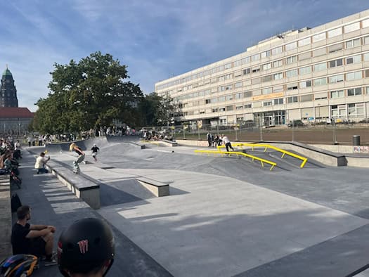 Sonniger Blick über den neuen Beton-Skatepark an der Lingnerallee in Dresden: Skateboarder fahren über graue Rampen und gelbe Geländer, am Rand schauen Besucher zu. Im Hintergrund steht ein Bürogebäude und der Rathausturm.