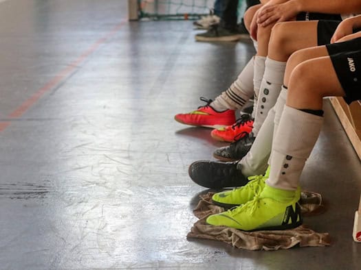 Kinder und Jugendliche sitzen nebeneinander auf einer Holzbank in einer Sporthalle. Die Nahaufnahme zeigt Beine in Sportkleidung, Stutzen und bunten Hallenschuhen auf dem Boden.