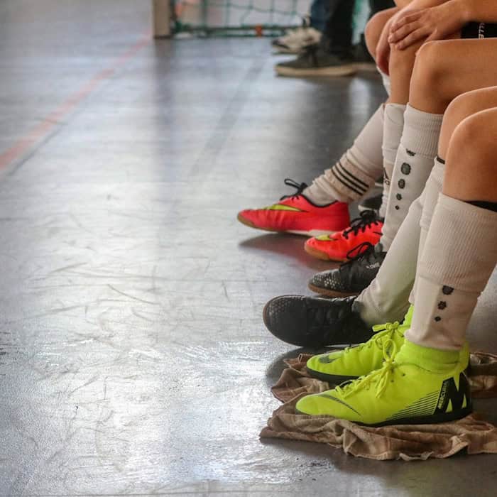 Kinder und Jugendliche sitzen nebeneinander auf einer Holzbank in einer Sporthalle. Die Nahaufnahme zeigt Beine in Sportkleidung, Stutzen und bunten Hallenschuhen auf dem Boden.