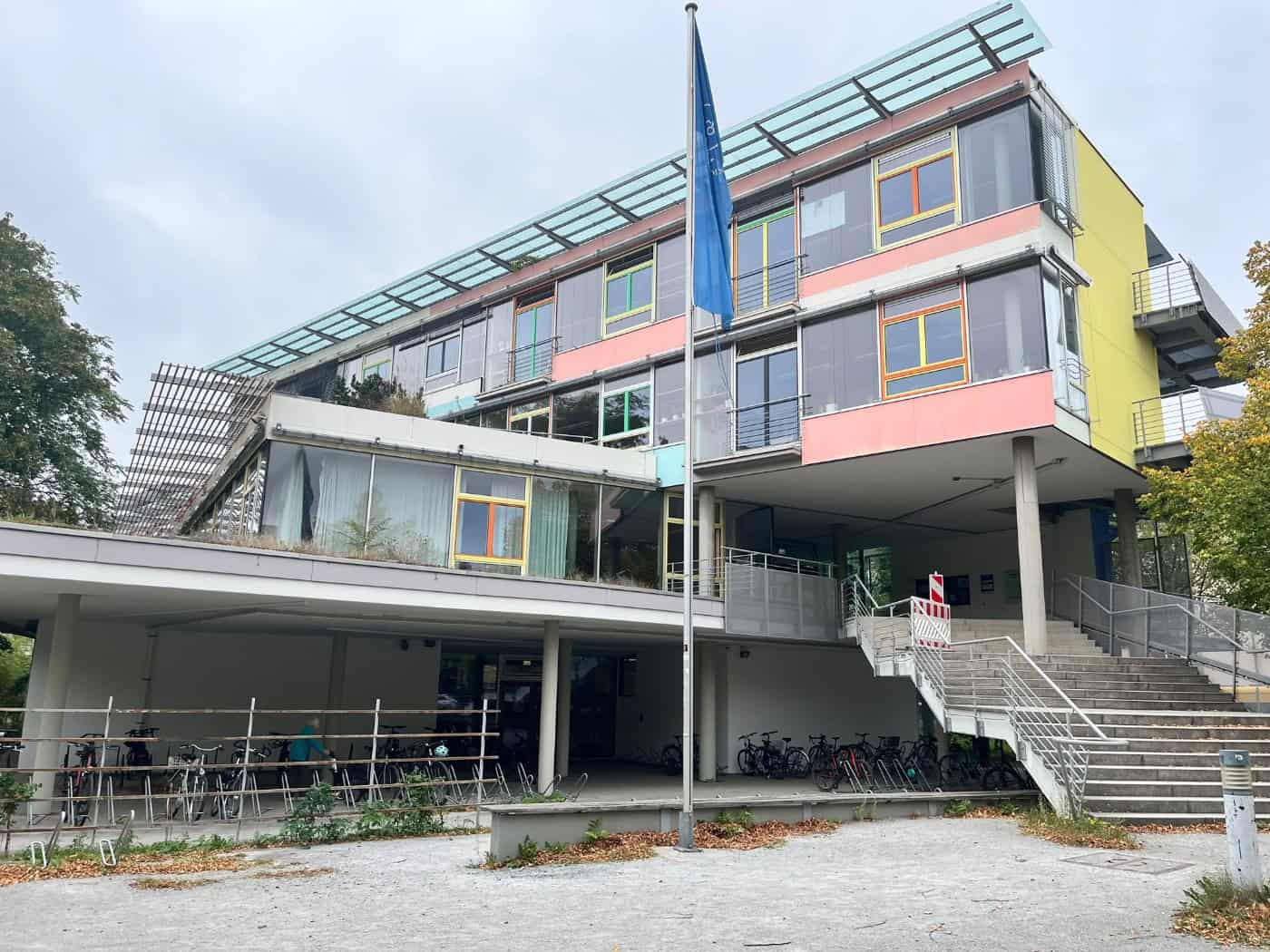 Außenansicht des St.-Benno-Gymnasiums in Dresden: Das moderne Schulgebäude hat eine bunte Fassade mit gelben, rosa und grünen Elementen, eine breite Außentreppe, Fahrradständer im Erdgeschoss und einen Fahnenmast im Vordergrund.