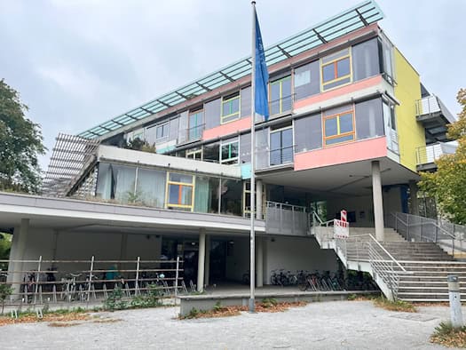 Außenansicht des St.-Benno-Gymnasiums in Dresden: Das moderne Schulgebäude hat eine bunte Fassade mit gelben, rosa und grünen Elementen, eine breite Außentreppe, Fahrradständer im Erdgeschoss und einen Fahnenmast im Vordergrund.